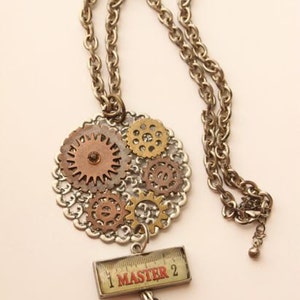 Puede incluir: Un collar con un colgante circular de engranajes metálicos en tonos bronce y oro sobre una base de filigrana plateada. Debajo cuelga un dije rectangular con un gráfico de regla y el texto rojo 1 MASTER 2, junto a un pequeño engranaje.