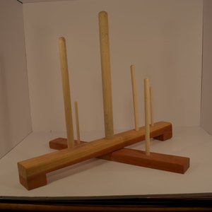 May include: A wooden stand with five dowels of varying lengths. The stand is made of two pieces of wood that are crossed at a right angle. The stand is a light brown color and the dowels are a natural wood color.