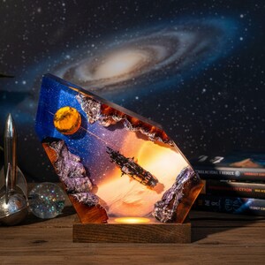 Könnte beinhalten: Eine Weltraum-Harzlampe mit einem Raumschiff-, Planeten- und Galaxie-Design. Die Lampe hat einen Holzsockel und ein warmes, leuchtendes Licht. Das Kunstwerk ist in klarem Harz eingeschlossen und erzeugt einen 3D-Effekt. Der Hintergrund zeigt eine Galaxie.