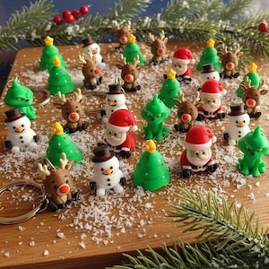 Pode incluir: Uma coleção de chaveiros com tema natalino, apresentando miniaturas de bonecos de neve, renas, árvores de Natal e figuras do Papai Noel. Os chaveiros são de várias cores, incluindo verde, vermelho, marrom e branco, e estão dispostos sobre uma superfície de madeira com neve artificial.