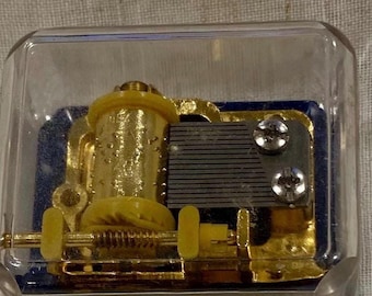 May include: A mechanical music box movement encased in a clear square plastic housing. The internal mechanism features a gold-colored rotating cylinder with pins, a silver-toned metal comb, and yellow plastic gears mounted on a blue base.