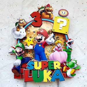 May include: A colorful Super Mario-themed cake topper featuring Mario, Luigi, Princess Peach, Yoshi, and other characters. The topper includes the name "Luka" and the number "3". The design is vibrant with red, blue, green, and yellow accents.