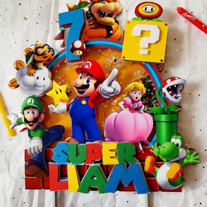 May include: A colorful Super Mario-themed cake topper featuring Mario, Luigi, Princess Peach, Yoshi, Bowser, and other characters. The topper includes the name "LIAM" and the number "7", with a "Happy Birthday" candle.