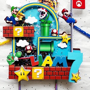 May include: A colorful cake topper featuring characters from the Super Mario Bros. video game. The design includes Mario, Luigi, Bowser, and other iconic elements like question mark blocks, stars, and a flag. The name "LIAM" and the number "7" are also included.