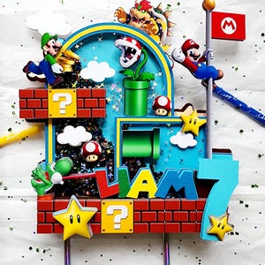 May include: A colorful birthday cake topper featuring characters from the Super Mario Bros. video game. The design includes Mario, Luigi, Bowser, Yoshi, and other iconic elements like question blocks, stars, and a flag. The name "LIAM" and the number "7" are also displayed.
