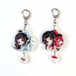 Könnte beinhalten: Zwei Acryl-Schlüsselanhänger mit Chibi-Figuren mit langem dunklem Haar. Die linke Figur trägt weiße Gewänder und hat einen kleinen Strohhut-Anhänger. Die rechte Figur trägt rote Gewänder und hat einen kleinen roten Regenschirm-Anhänger. Beide haben Metallverschlüsse.