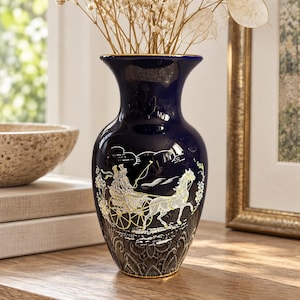 Könnte beinhalten: Eine tiefblaue Keramikvase mit goldfarbenem Rand an Öffnung und Boden. Der Körper zeigt eine detaillierte gold-weiße Illustration einer Pferdekutsche mit zwei Figuren vor einem strukturierten, dunkelgrauen spitzenartigen Muster.