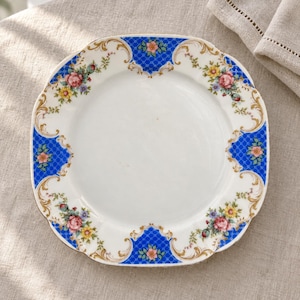 Può includere: Un piatto quadrato in porcellana con angoli arrotondati, centro bianco e bordo decorativo. Il bordo presenta un motivo a griglia blu, volute dorate e grappoli di motivi floreali rosa, gialli e verdi lungo i bordi del piatto.
