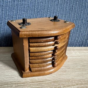 Könnte beinhalten: Ein hölzerner Untersetzerhalter mit einem Stapel aus sechs geschwungenen Holzuntersetzern. Der Halter hat eine glatte Oberfläche mit zwei dunklen vertikalen Stiften und vier kleinen dekorativen silberfarbenen Metallnieten an den Ecken.