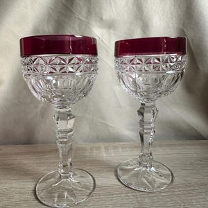 May include: Two clear crystal wine glasses with a ruby red rim. The glasses have a detailed, cut-glass design on the bowl and a faceted stem. The glasses are displayed on a light-colored wooden surface.