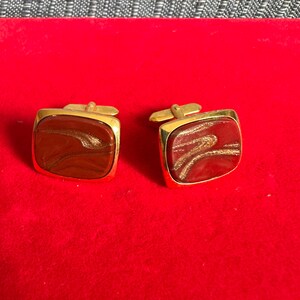 Peut inclure: Une paire de boutons de manchette rectangulaires dorés avec une incrustation marbrée rouge foncé et marron, ornée de fines lignes tourbillonnantes dorées et scintillantes. Chaque bouton possède un cadre en métal doré et une tige pivotante.