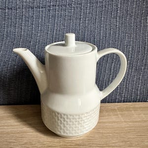 Könnte beinhalten: Eine weiße Teekanne aus Keramik mit einem glatten oberen Bereich und einem strukturierten, geometrischen Wabenmuster am unteren Sockel. Die Kanne verfügt über einen zylindrischen Körper, einen geschwungenen Griff, einen Ausguss und einen Deckel.