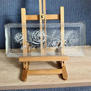 Op de afbeelding: Een rechthoekig dienblad van helder glas met een geëtst wit bloemmotief van rozen en bladeren. Het dienblad heeft een decoratieve rand met herhalende verticale lijnen en wordt gepresenteerd op een houten schildersezel.