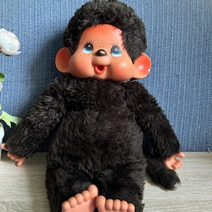 Pode incluir: Uma boneca de pelúcia em forma de macaco com pelo castanho escuro, rosto de vinil cor de pêssego, olhos grandes azuis e nariz pequeno. A boneca apresenta orelhas proeminentes, boca aberta, sardas no nariz e mãos e pés cor de pêssego a condizer.