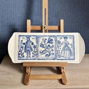 May include: A rectangular off-white ceramic serving tray featuring three blue illustrations. The left panel shows a person holding a glass with the text 17 and 70. The centre panel displays a floral motif. The right panel depicts a person with the text H and H.