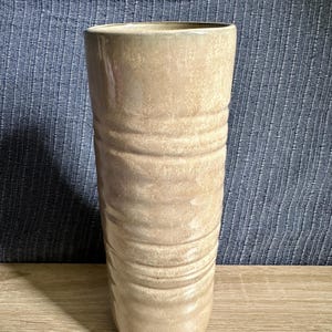 Könnte beinhalten: Eine hohe, zylindrische Keramikvase mit einer hellbeigen, gesprenkelten Glasur. Die Außenfläche weist eine Reihe horizontaler, wellenförmiger Rillen auf, die sich um den Körper ziehen und vom Rand bis zum Boden ein strukturiertes Aussehen verleihen.