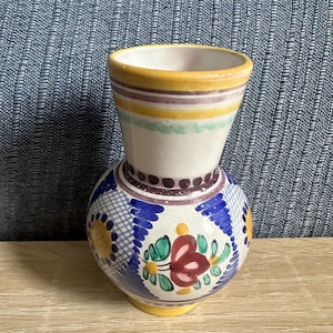 May include: A ceramic vase with a white base, featuring a hand-painted design of blue cross-hatching, yellow circular accents, and a central floral motif in red and green. The rim is painted yellow, with purple and green horizontal bands.