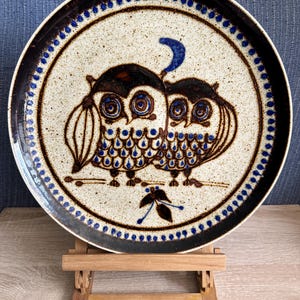 Op de afbeelding: Een rond keramieken bord met een gespikkelde crèmekleurige basis met een donkerbruine en blauwe illustratie van twee uilen naast elkaar. Een blauwe maansikkel bevindt zich boven de uilen en een klein donkerbruin bladmotief is eronder gecentreerd.