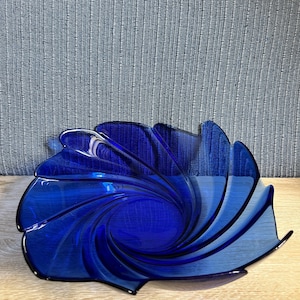May include: A deep cobalt blue glass bowl featuring a distinct spiral pattern that radiates from the center toward the scalloped, petal-like edges. The bowl is constructed from translucent, smooth glass with a glossy finish and a curved, open form.