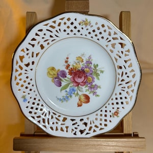 Puede incluir: Plato de porcelana blanca con borde calado y borde dorado. El centro del plato presenta un diseño floral con flores rojas, moradas, amarillas y azules. El plato se exhibe sobre un caballete de madera.