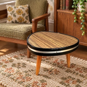 Puede incluir: Una mesa de centro triangular en miniatura con una superficie de veta de madera, un borde negro y una banda plateada delgada. La mesa cuenta con tres patas de madera clara que se estrechan hacia abajo, proporcionando una base estable para este mueble.