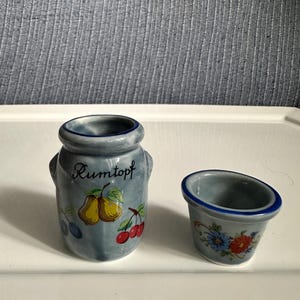 May include: A small ceramic jar with a blue-grey glaze and the word Rumtopf printed in black script. The jar features painted yellow pears, red cherries, and blue plums. Beside it sits a matching small ceramic bowl with blue floral accents.