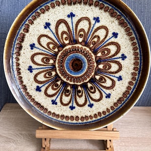 May include: A round ceramic plate featuring a speckled cream-colored base with a central brown and blue floral-inspired pattern. The design consists of concentric circles and radiating petals, finished with a dark brown rim and a decorative border.