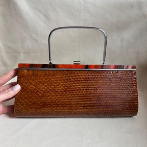 May include: A vintage brown handbag with a reptile skin pattern. The bag has a silver handle and a tortoiseshell-patterned top with orange accents. The rectangular bag is held by a hand.