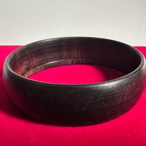 Op de afbeelding: Een ronde, donkerbruine houten kom met een gladde, gepolijste buitenkant en een zichtbaar natuurlijk houtnerfpatroon. De kom heeft een dikke, afgeronde rand en een diepe, holle binnenkant die de donkere textuur van het houtmateriaal toont.