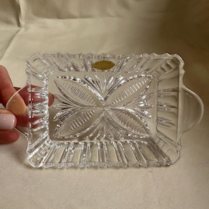 May include: Clear rectangular crystal dish with a floral design in the center and a textured border. The dish has small handles on each side. A small gold label is visible.