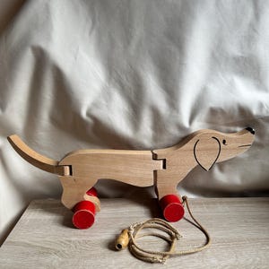 Könnte beinhalten: Ein hölzernes Dackel-Spielzeug zum Ziehen mit roten Rädern und einer Schnur. Der Hund ist aus hellem Holz mit schwarzen Details für Augen und Nase gefertigt. Das Spielzeug ist mit Gelenken für Bewegung ausgestattet.