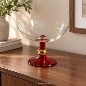Può includere: Una ciotola in vetro trasparente con un bacino ampio e poco profondo sostenuto da una base a piedistallo. Lo stelo presenta una sezione in vetro rosso intenso, un accento sferico color oro lucido e un piede circolare in vetro rosso.