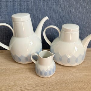 Peut inclure: Deux théières en porcelaine blanche avec couvercles et un pot à lait assorti. Chaque pièce présente un corps arrondi, une anse incurvée, un long bec verseur et un motif répétitif de pétales bleu clair autour de la base des récipients.
