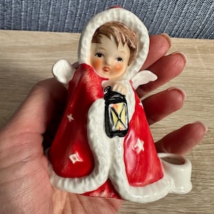 Puede incluir: Una figura de cerámica de un niño con cabello castaño que viste una capa roja con capucha y borde de piel blanca. El niño sostiene una linterna negra con luz amarilla. La capa tiene estrellas blancas y un pequeño portavelas circular adjunto.