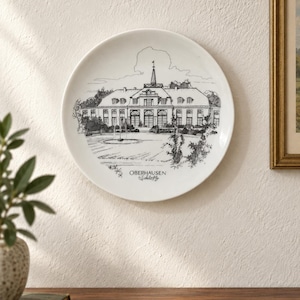 May include: A round white ceramic decorative plate featuring a detailed black line drawing of the Oberhausen Schloss building, including a central fountain and surrounding trees. The text OBERHAUSEN Schloss is printed in black below the illustration.