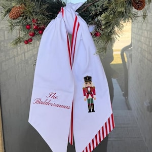 Puede incluir: Una corona navideña con agujas de pino, bayas rojas y piñas. Un tejido blanco con rayas rojas y un diseño de cascanueces cuelga de la corona. El texto "The Balderramas" está bordado en la tela.