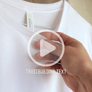 May include: A close-up of a white t-shirt hanging on a metal hanger. The shirt has a tag that reads "Canvas Bella-Canvas". The text "TRUSTBUILDING TEXT" is displayed on the shirt.