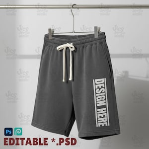 Może przedstawiać: Szorty dresowe w kolorze szarym z białym sznurkiem i napisem "DESIGN HERE" wydrukowanym pionowo na nogawce. Szorty wiszą na metalowym wieszaku na neutralnym tle. Tekst "EDITABLE *.PSD" znajduje się na dole.