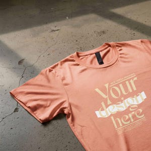 May include: A peach-coloured short-sleeve t-shirt with a crew neckline and a black inner collar tag. The front features the text "VICE VERSA MOCKUPS" above the words "Your design here" printed in a light cream colour with a white horizontal stripe.