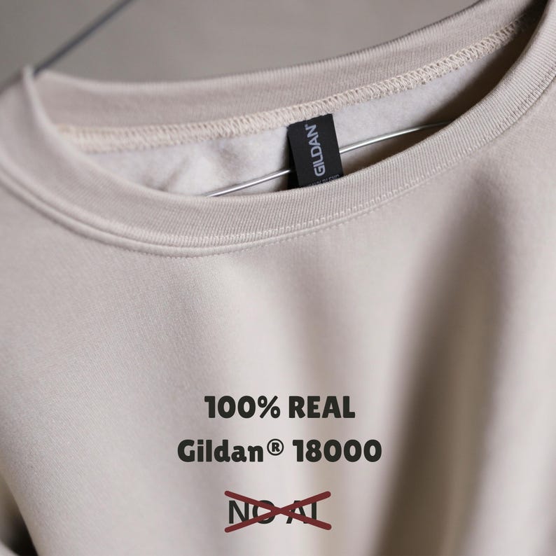 Puede incluir: Sudadera beige Gildan 18000 con el texto "100% REAL" y "NO AI" tachado en rojo. La sudadera est&aacute; colgada en una percha met&aacute;lica.