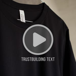 May include: Black t-shirt with a white play button graphic and the text "TRUSTBUILDING TEXT". The shirt has a crew neck and a tag that reads "CANVAS BELLA+CANVAS".