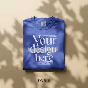 May include: A folded Flo Blue t-shirt with the text "Your design here" in white. The shirt is a mock-up, with the words "VICE VERSA MOCKUPS" above the text and a small tag on the collar. The shirt is on a neutral background.