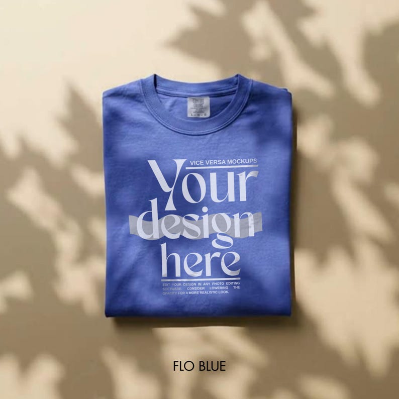 May include: A folded Flo Blue t-shirt with the text "Your design here" in white. The shirt is a mock-up, with the words "VICE VERSA MOCKUPS" above the text and a small tag on the collar. The shirt is on a neutral background.