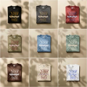 May include: Nine folded t-shirts in various colors, including black, gray, red, olive green, blue, and white. Each shirt has the text "Your design here" printed on the front in a contrasting color. The shirts are arranged on a neutral background.