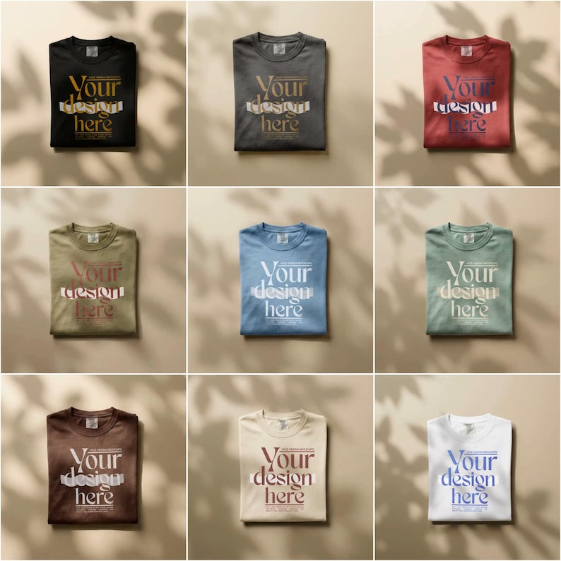 May include: Nine folded t-shirts in various colors, including black, gray, red, olive green, blue, and white. Each shirt has the text "Your design here" printed on the front in a contrasting color. The shirts are arranged on a neutral background.