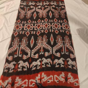 Puede incluir: Textil rectangular con fondo negro, con patrones geométricos y animales rojos y blancos. El textil tiene un borde con flecos y es probablemente una bufanda o chal.
