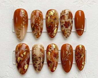 May include: A set of ten almond-shaped press-on nails featuring a warm autumn color palette of burnt orange, cream, and brown. The designs include gold leaf accents, hand-painted maple leaves, delicate vine illustrations, and a forest scene.
