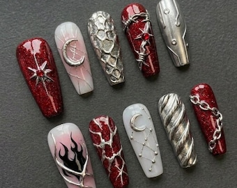 May include: A set of ten press-on nails featuring a mix of deep red glitter, white, and metallic silver finishes. Designs include silver chains, crescent moons, barbed wire, flame motifs, and textured patterns with small red gemstone accents.