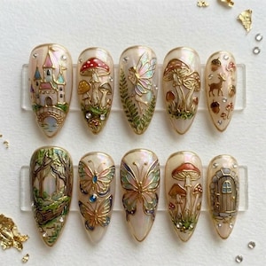 May include: A set of ten almond-shaped press-on nails featuring intricate 3D designs including a castle, mushrooms, a fairy, a deer, butterflies, a forest scene, and a wooden door, all accented with gold metallic details and small clear rhinestones.