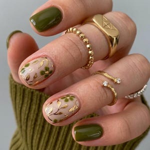 May include: A set of gold rings worn on fingers with manicured nails. Two nails feature a solid olive green polish, while two nails display a nude base with gold leaf floral patterns. The rings include a signet ring, a beaded band, and open bands.